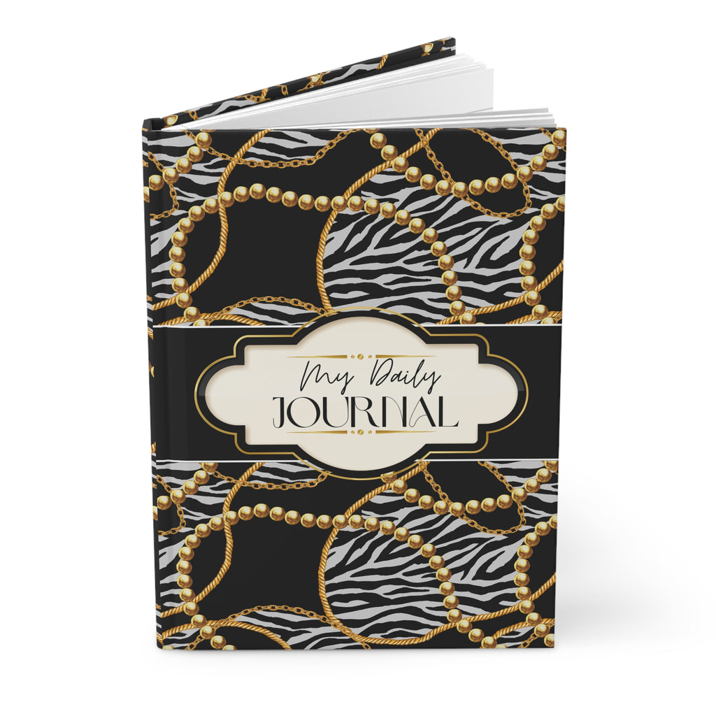 A hardcover daily journal with a black and white zebra print cover decorated with gold chain and pearl patterns, featuring a central cream-colored label that reads 'My Daily JOURNAL'.
