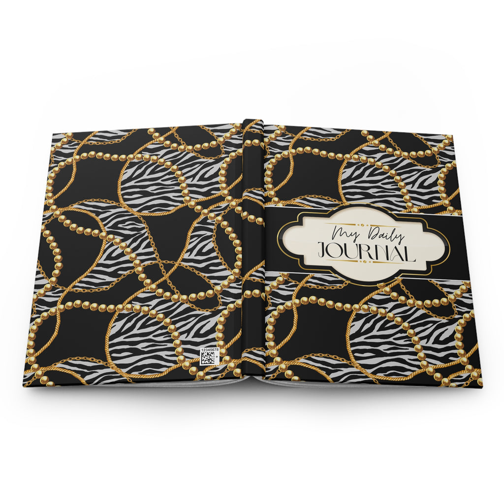 An open daily journal with a black and white zebra print cover patterned with gold chains and gold beads, featuring a cream label on the right page that reads 'My Daily JOURNAL'.