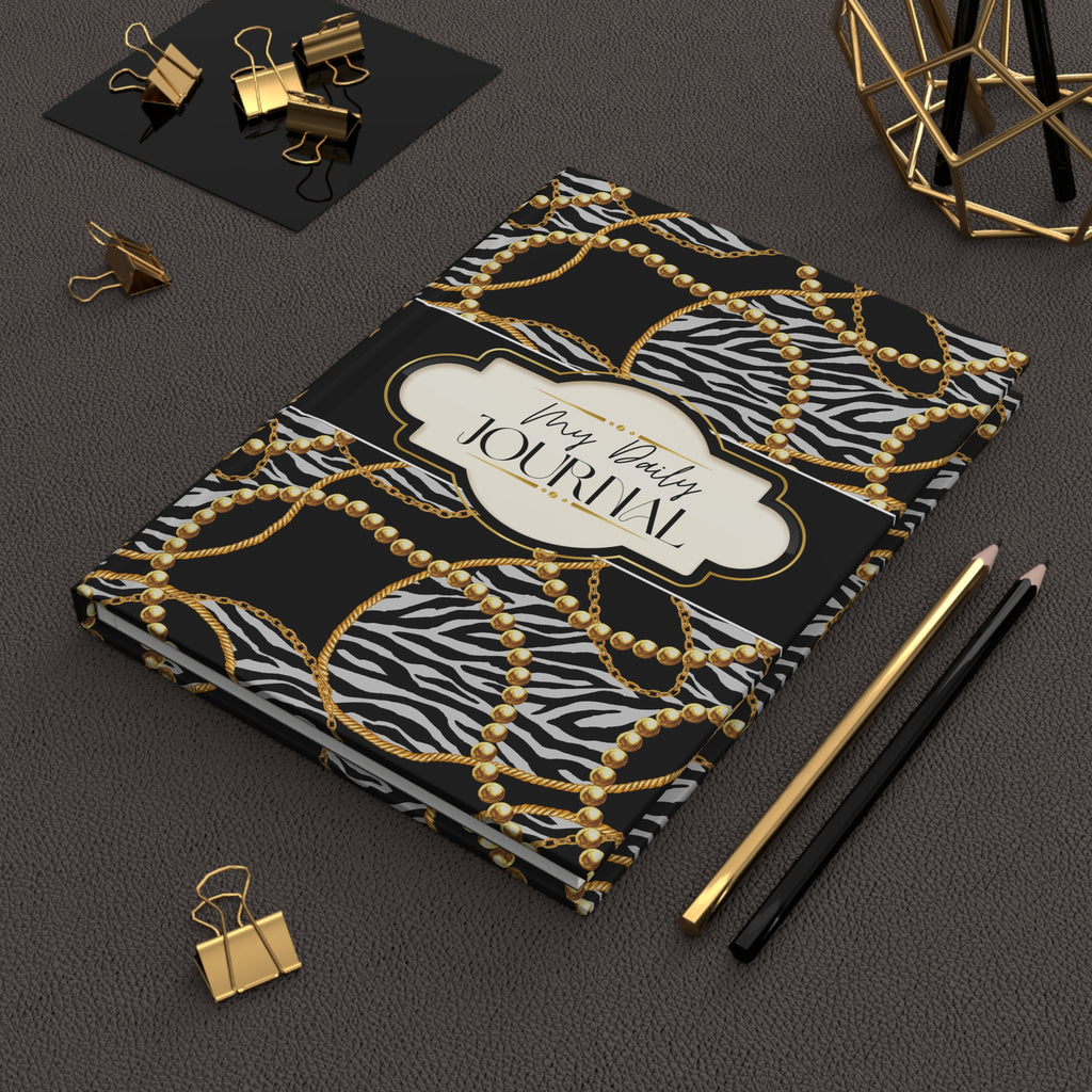 A hardcover journal with a black and white zebra print background, overlaid with a gold chain and pearl pattern, and a cream label reading 'My Daily Journal'. The journal is styled on a dark textured surface with gold and black office accessories.