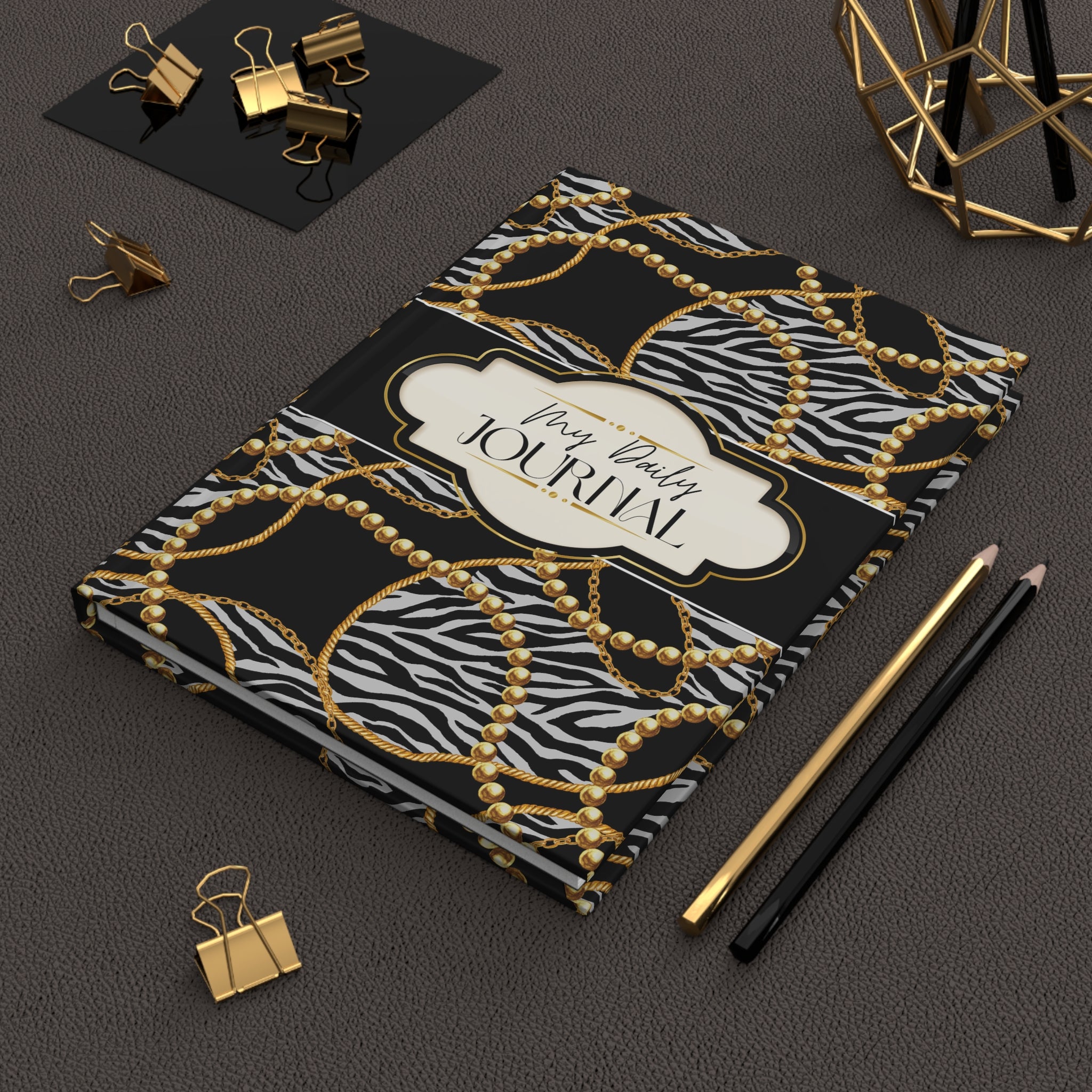 A hardcover journal with a black and white zebra print background, overlaid with a gold chain and pearl pattern, and a cream label reading 'My Daily Journal'. The journal is styled on a dark textured surface with gold and black office accessories.