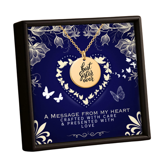 A gold round pendant necklace engraved with 'best sister ever' hangs from a gold chain inside a square dark navy gift box. The box features a decorative navy and white floral heart design with the text 'A MESSAGE FROM MY HEART CRAFTED WITH CARE & PRESENTED WITH LOVE'.
