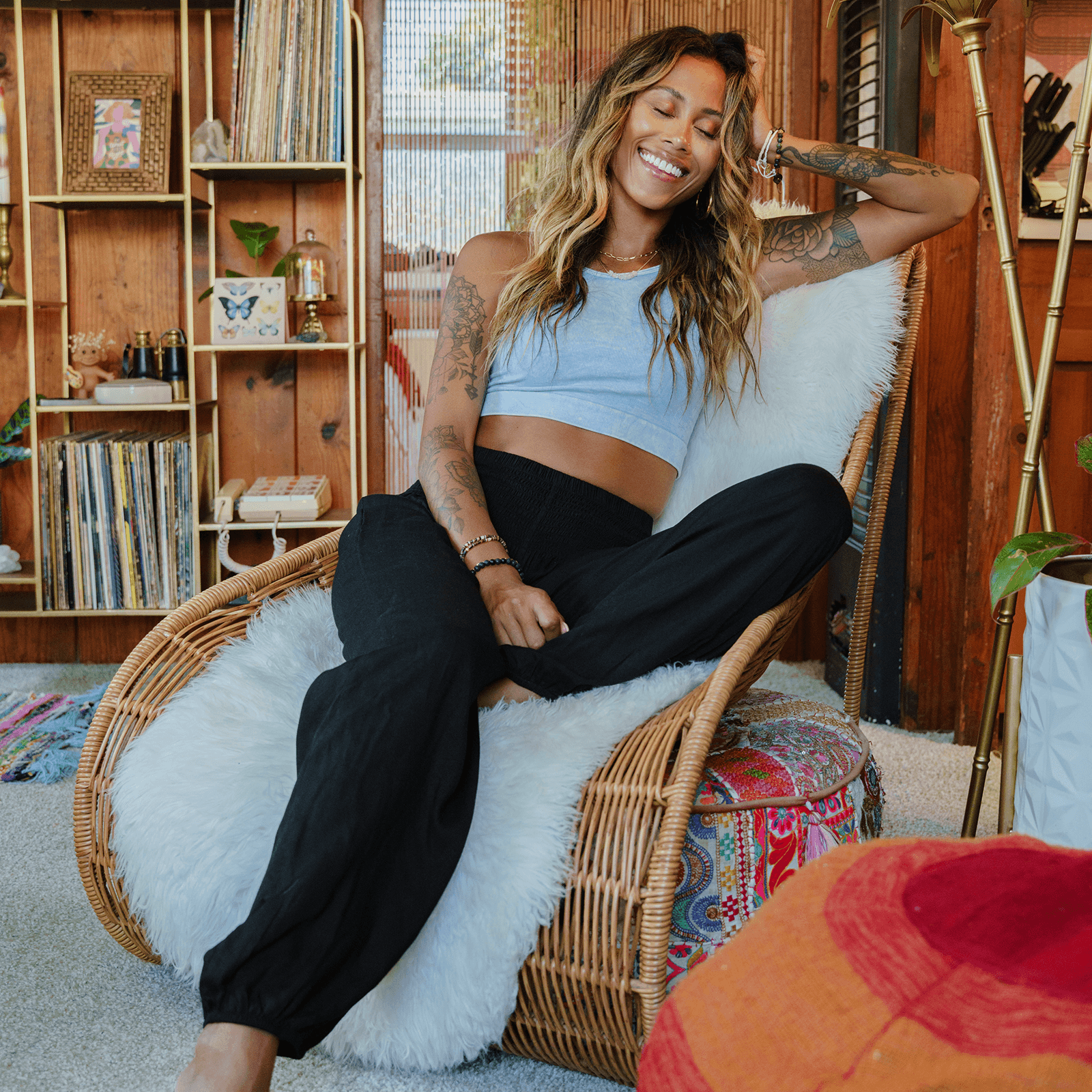 Woman sitting on a wicker chair in a cozy room with wooden shelves and plants.
