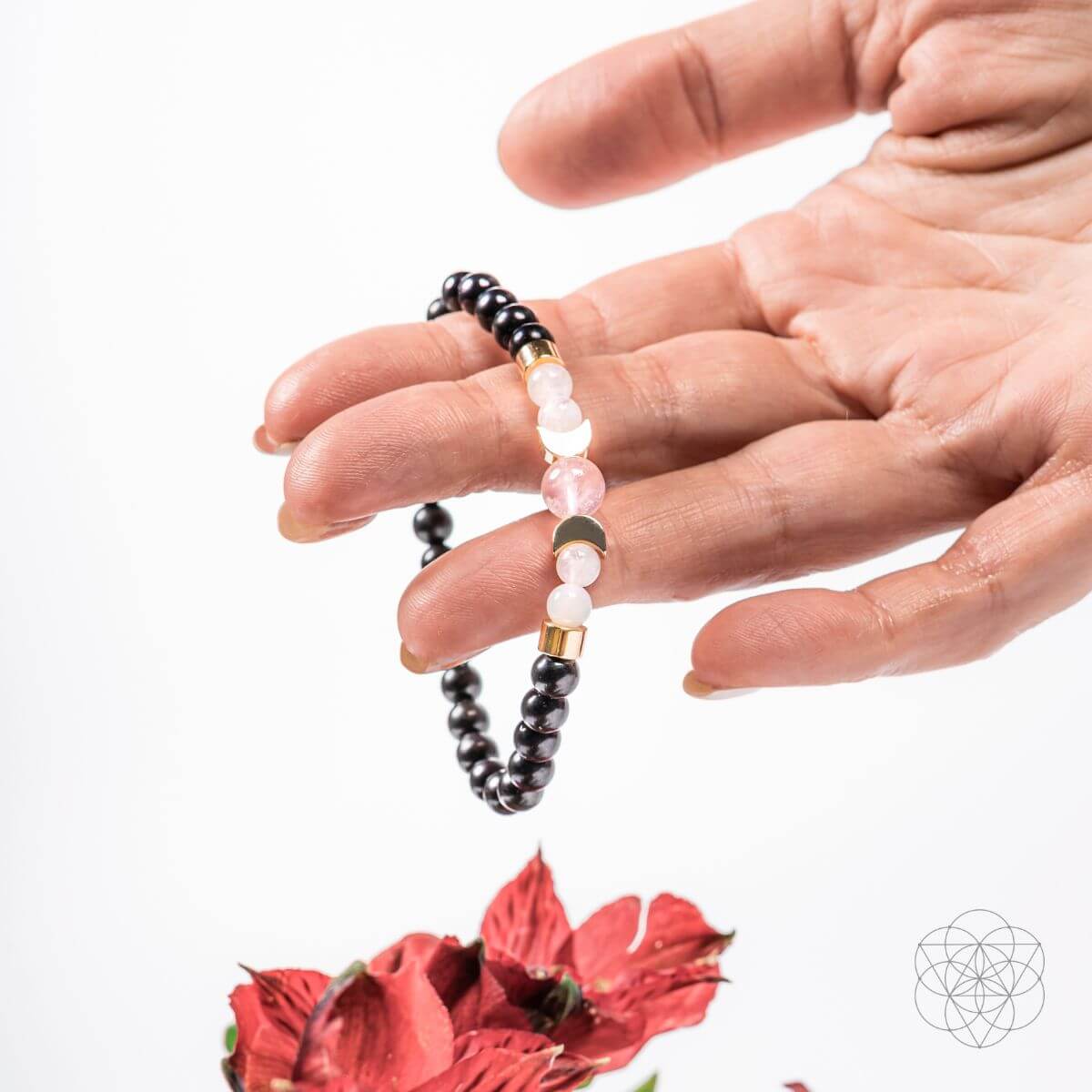 Hand holding a beaded bracelet with black, white, and gold beads on a white background with a red flower.