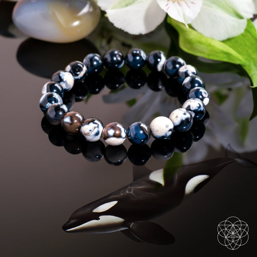 Beaded bracelet with black, blue, and silver beads on a reflective surface with flowers in the background