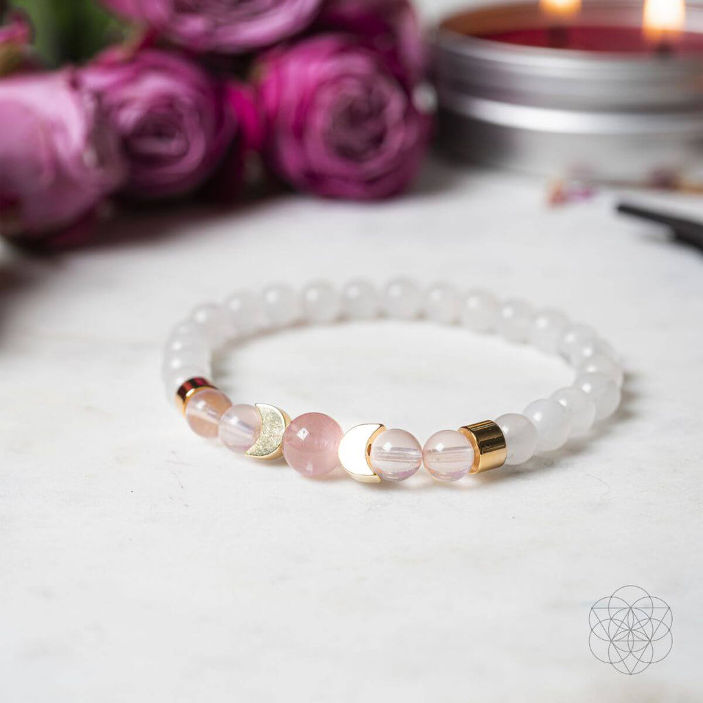 Bracelet with pink and gold beads on a white surface with purple flowers and candles in the background