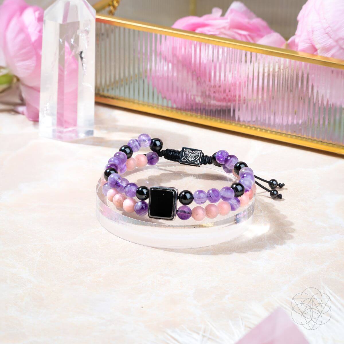 Stack of beaded bracelets with a black square charm on a white surface with pink flowers and crystals in the background.