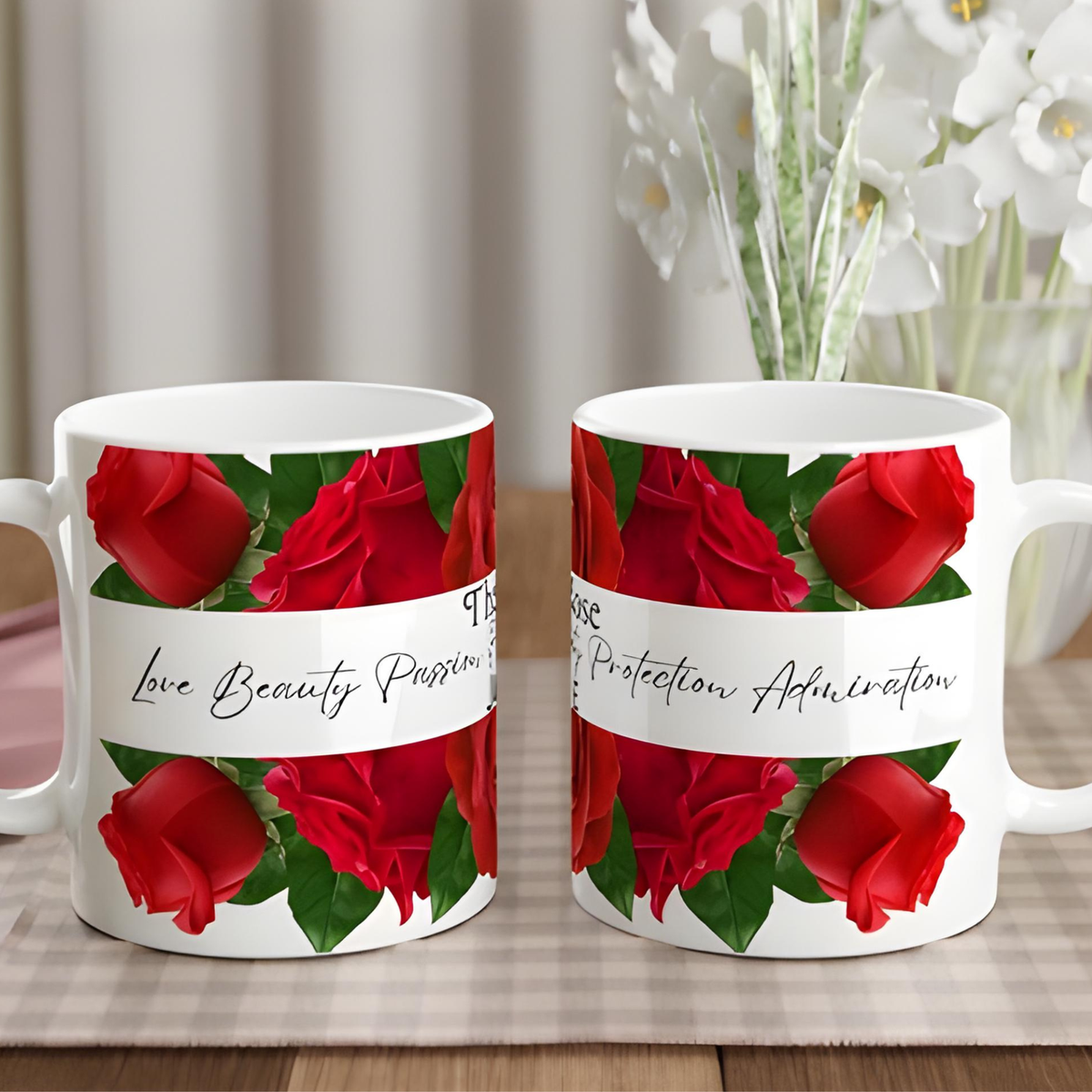 Two white ceramic mugs with a red rose and green leaf pattern, featuring a white banner with black cursive text, displayed on a table with a blurred background of flowers.