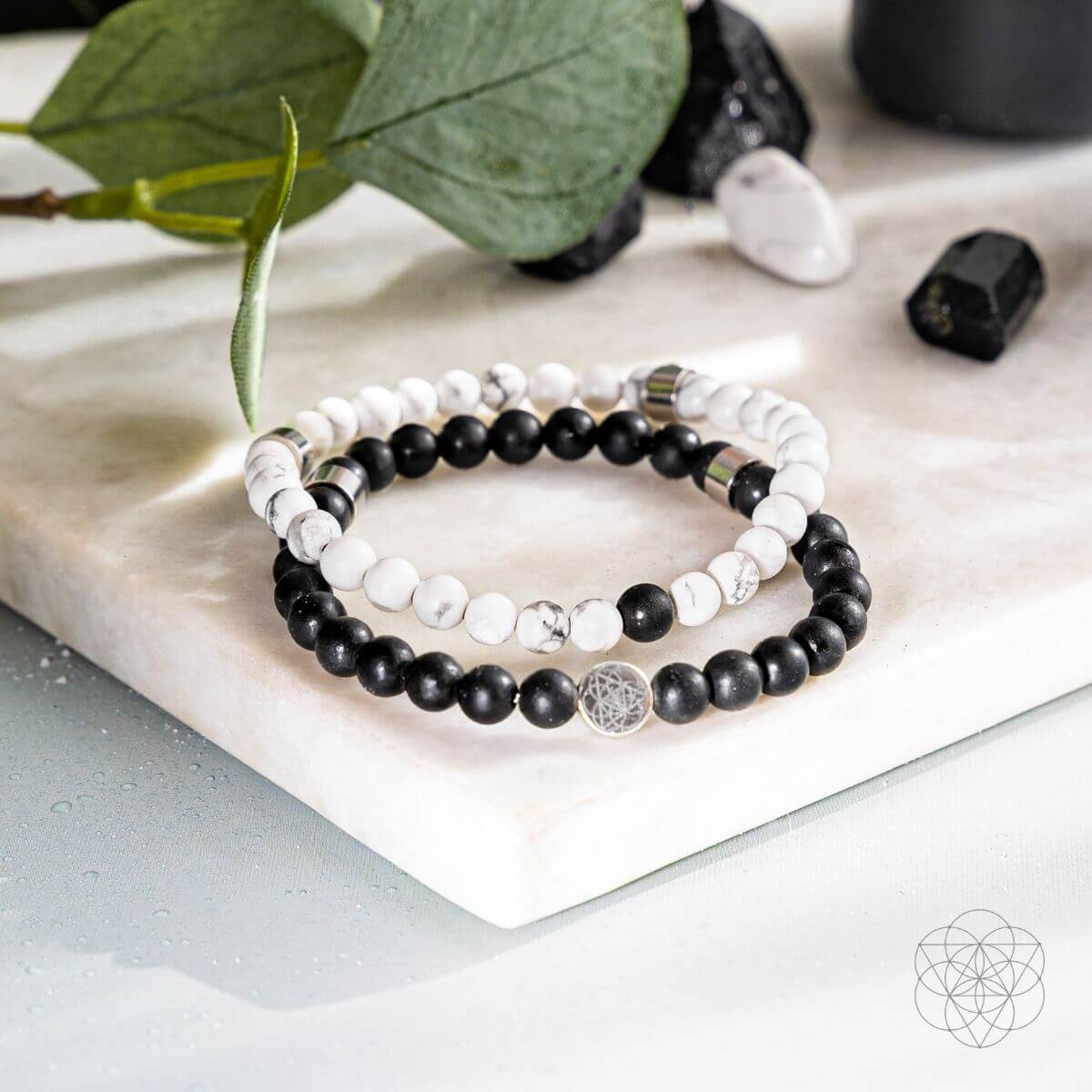 Stack of black and white beaded bracelets on a marble surface with green leaves and stones.