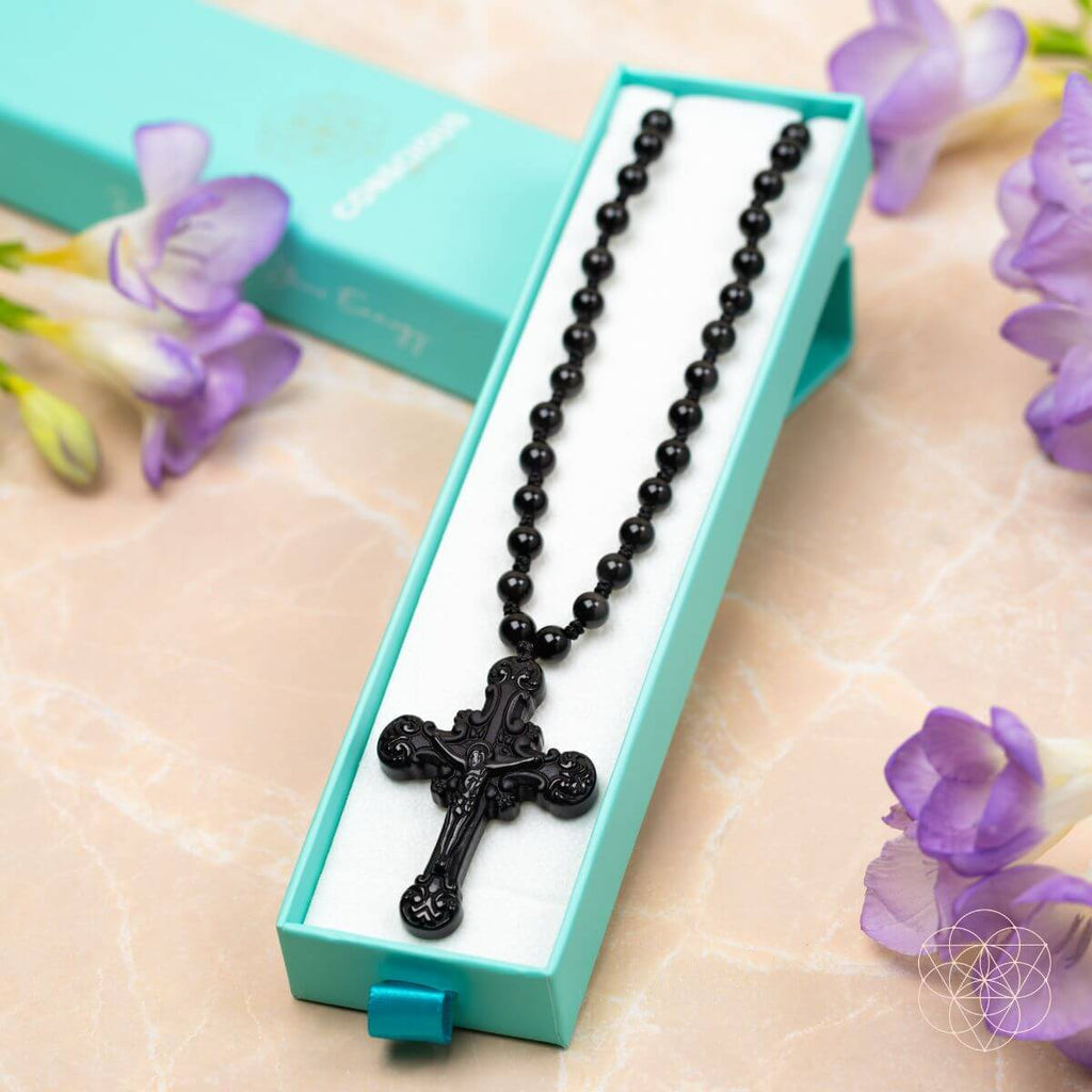 Black beaded necklace with a cross pendant in a turquoise box, surrounded by purple flowers.