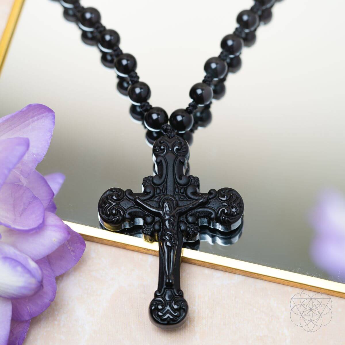 Black beaded necklace with an ornate cross pendant on a reflective surface with purple flowers.