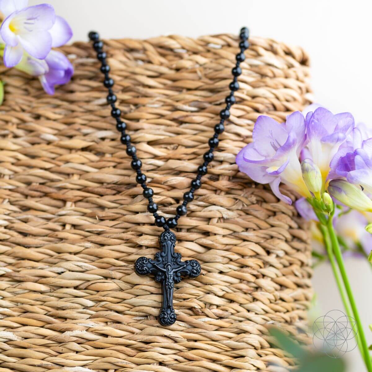 Black beaded necklace with a cross pendant on a woven surface with purple flowers.