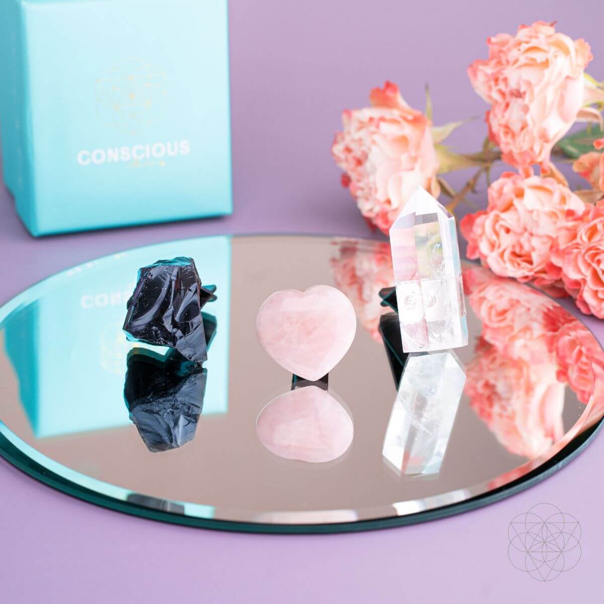 Mirror with crystals on a purple surface, next to a turquoise box and pink flowers.