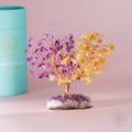 Crystal tree with purple and yellow stones next to a teal cylindrical container on a pink background