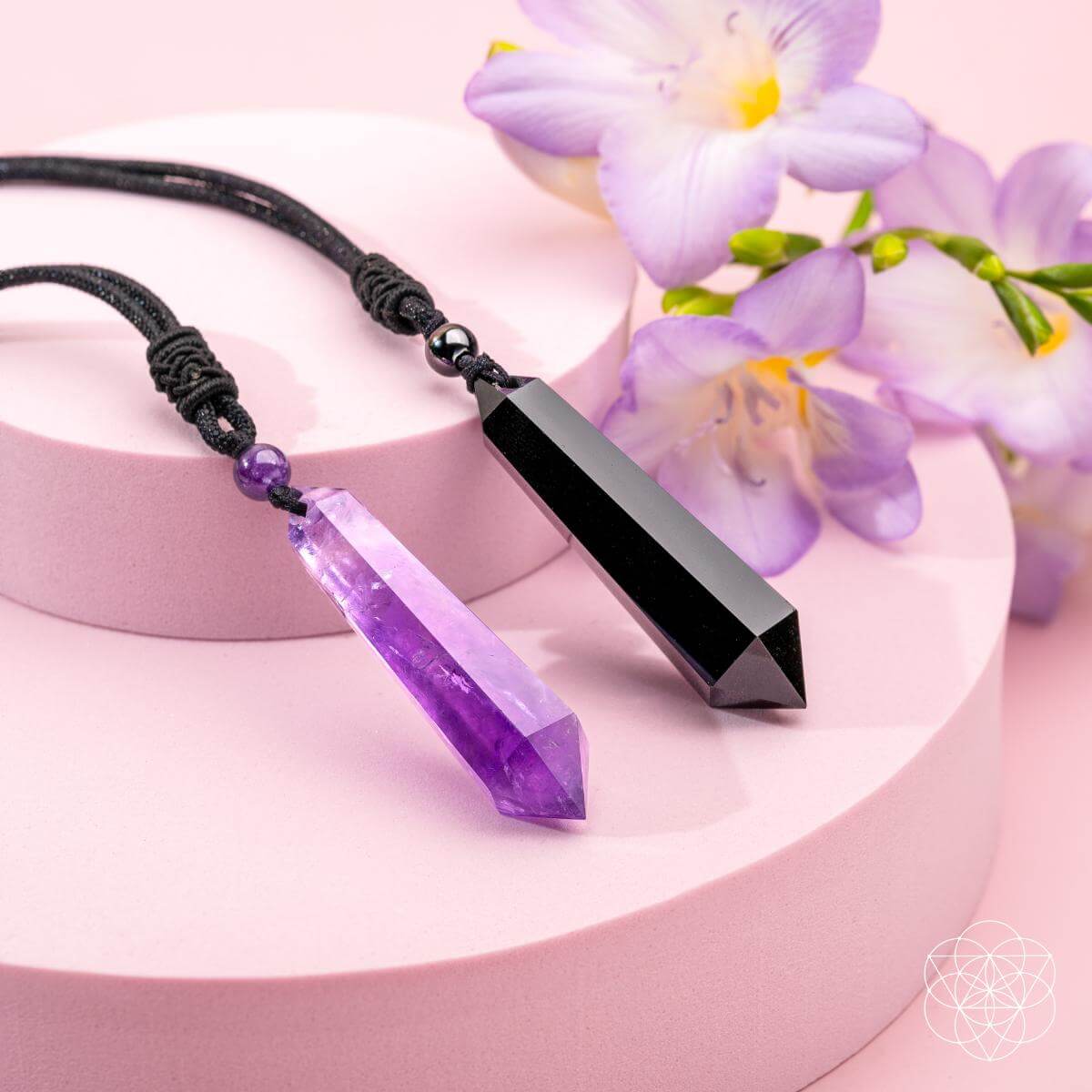 Necklace with purple crystal pendant and black gemstone on a pink surface with flowers.