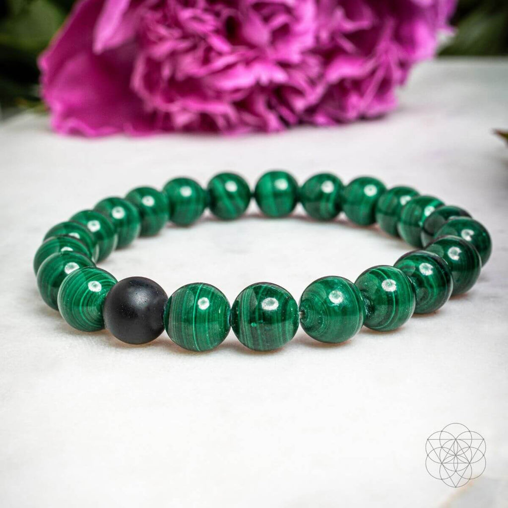 Green beaded bracelet with a black bead on a white surface with pink flowers in the background