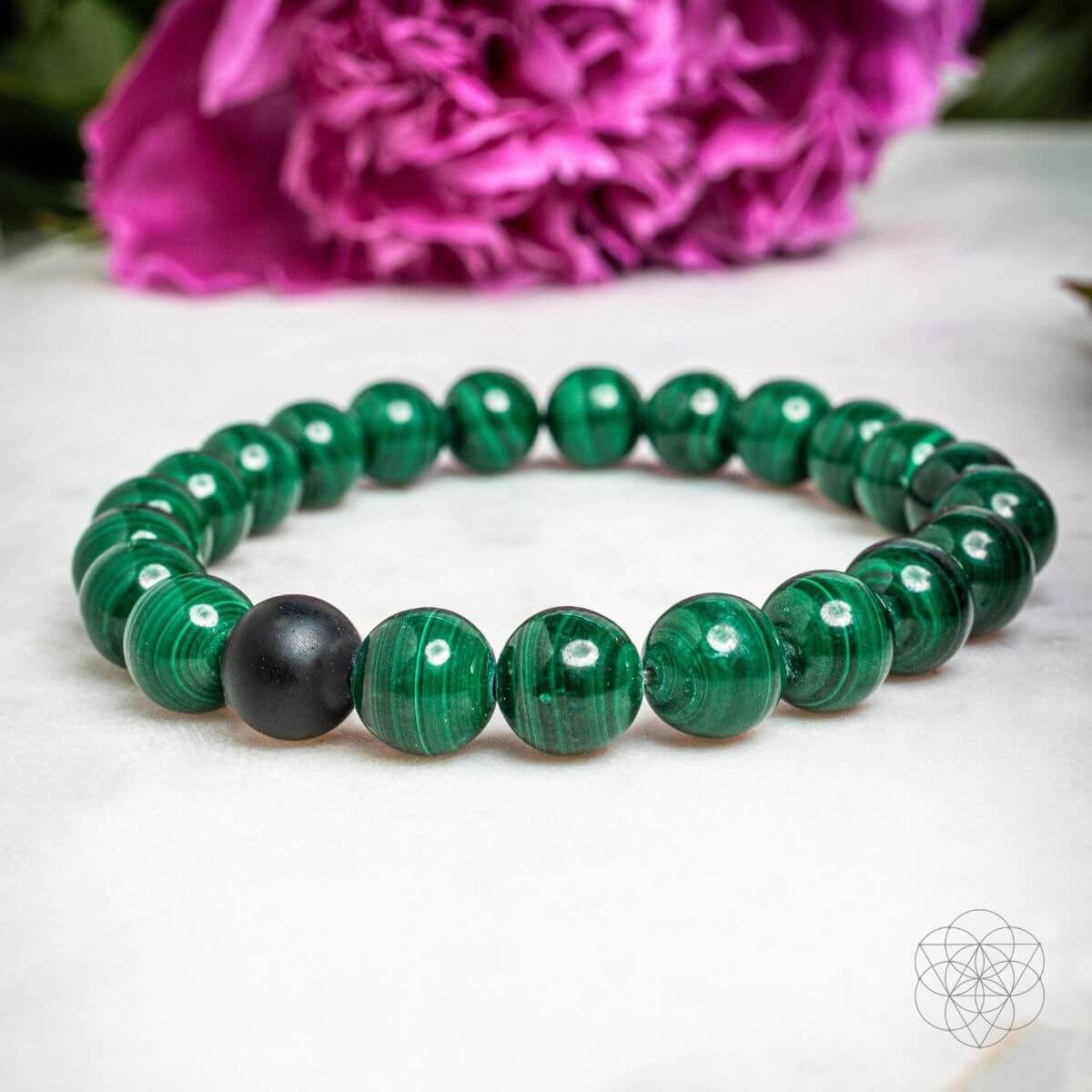 Green beaded bracelet with a black bead on a white surface with pink flowers in the background