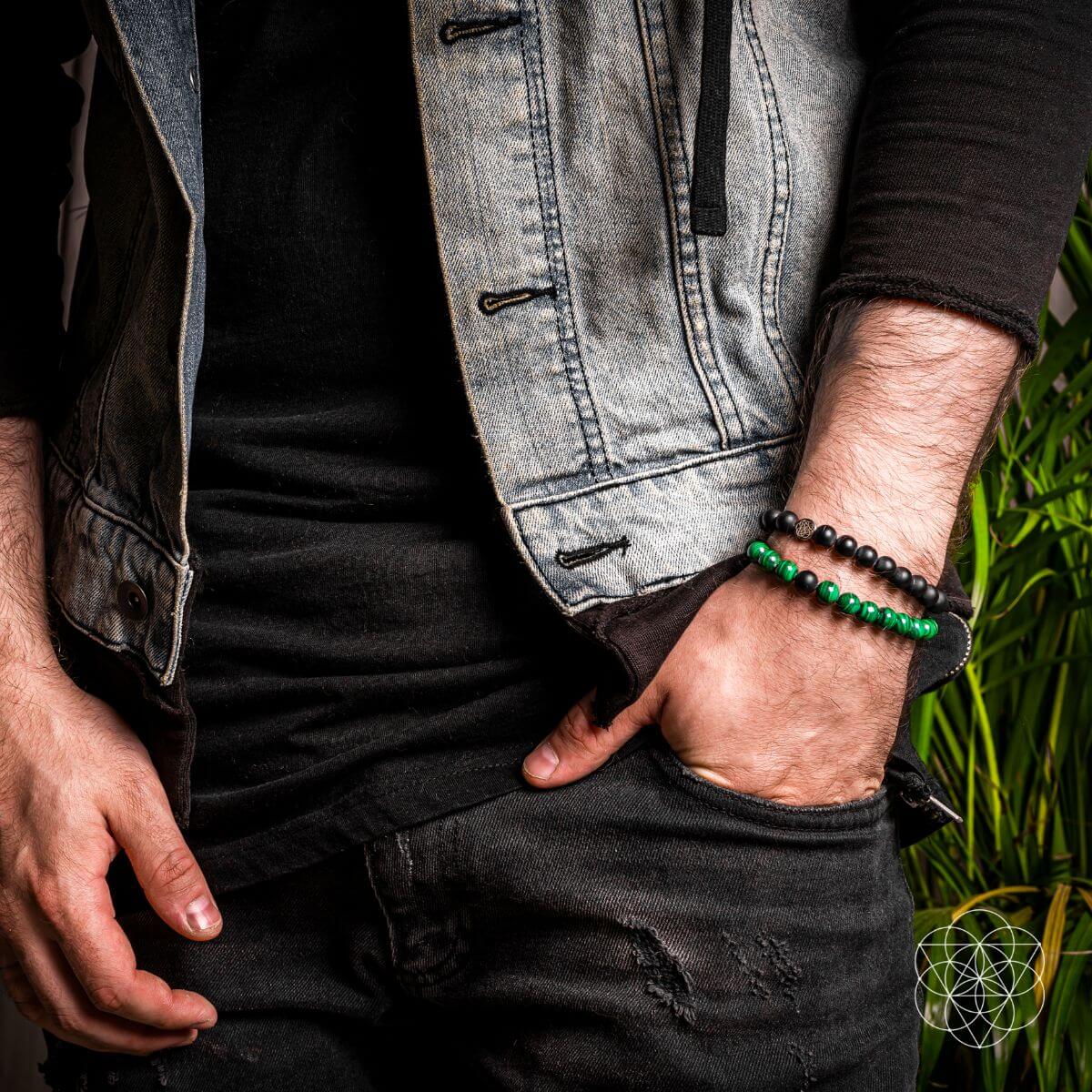 Person wearing a denim jacket and black shirt with green and black bracelets on a blurred natural background