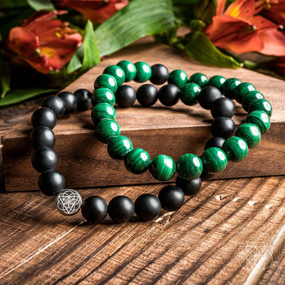Two green and black beaded bracelets on a wooden surface with flowers in the background.