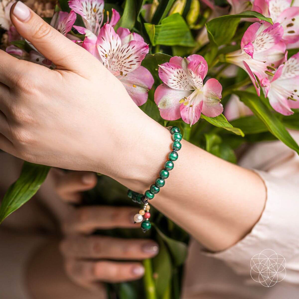 Hand wearing a green beaded bracelet with pink flowers in the background