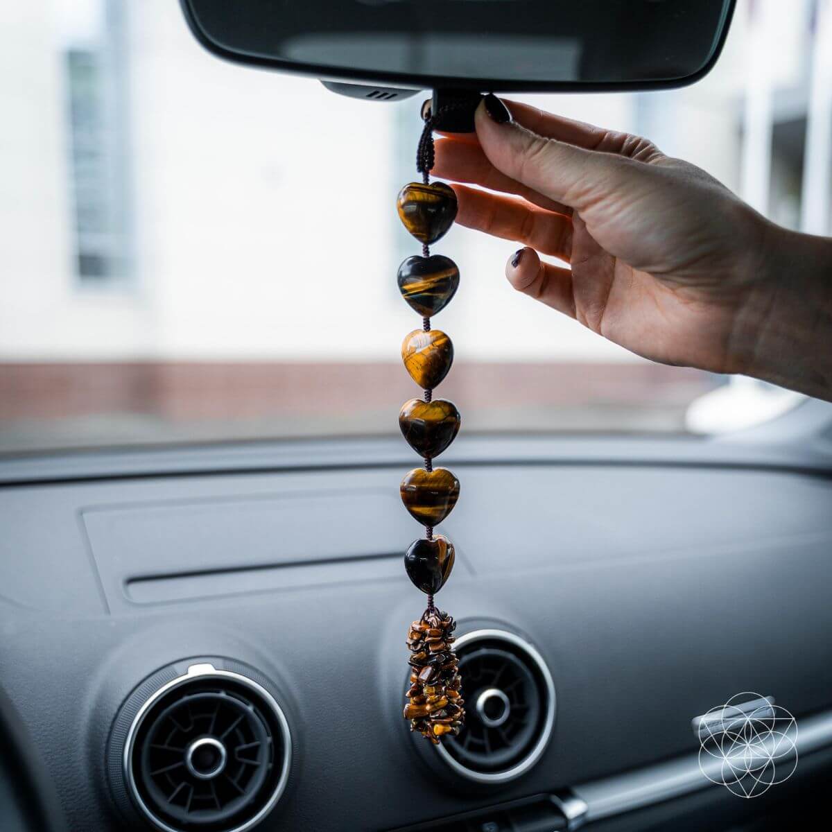 Hand holding a beaded car hanging charm in front of a car's rearview mirror.