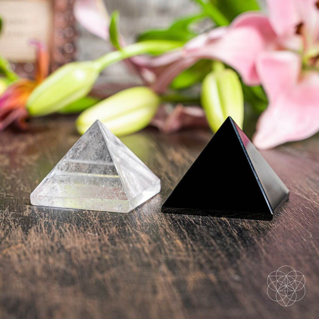 Two pyramid-shaped crystals on a wooden surface with flowers in the background