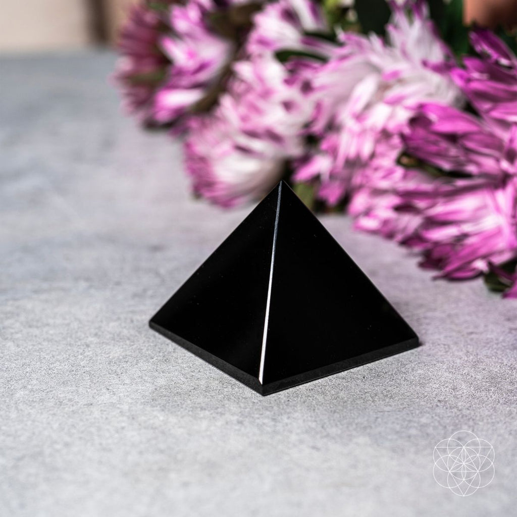 Black pyramid crystal on a gray surface with pink flowers in the background