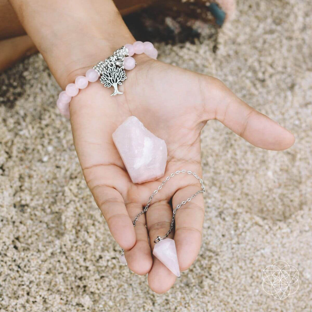 Hand holding a pink crystal with a bracelet featuring a tree of life charm on a sandy background