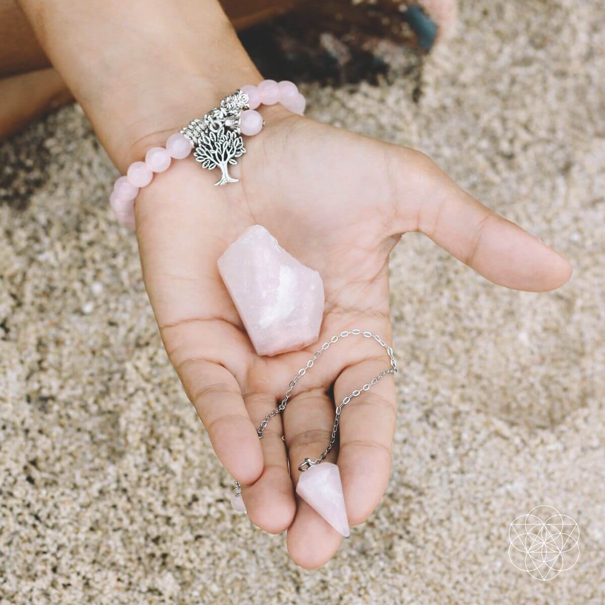 Hand holding a pink crystal with a bracelet featuring a tree of life charm on a sandy background