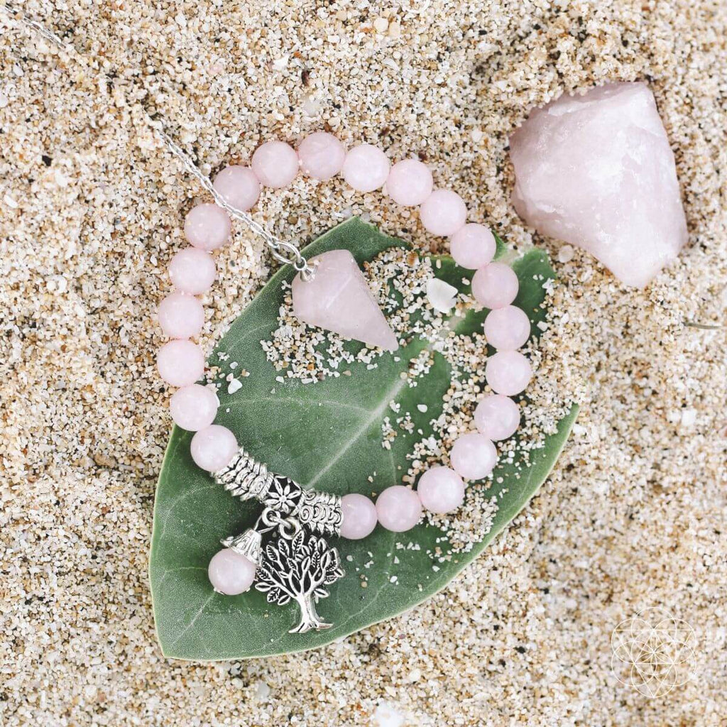 Pink beaded bracelet with a silver tree charm on a green leaf, lying on sandy surface.