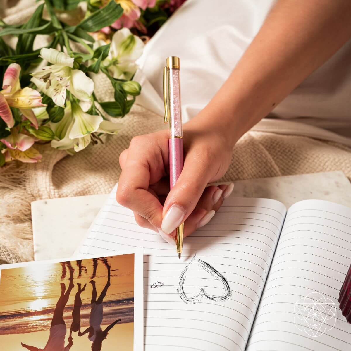 Hand holding a pink pen over an open notebook with a heart drawing, next to a bouquet of flowers and a photo of a sunset.