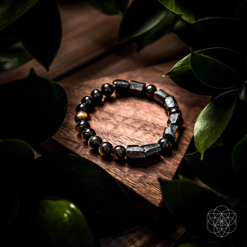 A beaded stretch bracelet made of polished black and metallic silver beads with a few gold-toned accents, displayed on a wooden block surrounded by dark green leaves.