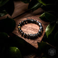 A beaded stretch bracelet made of polished black and metallic silver beads with a few gold-toned accents, displayed on a wooden block surrounded by dark green leaves.