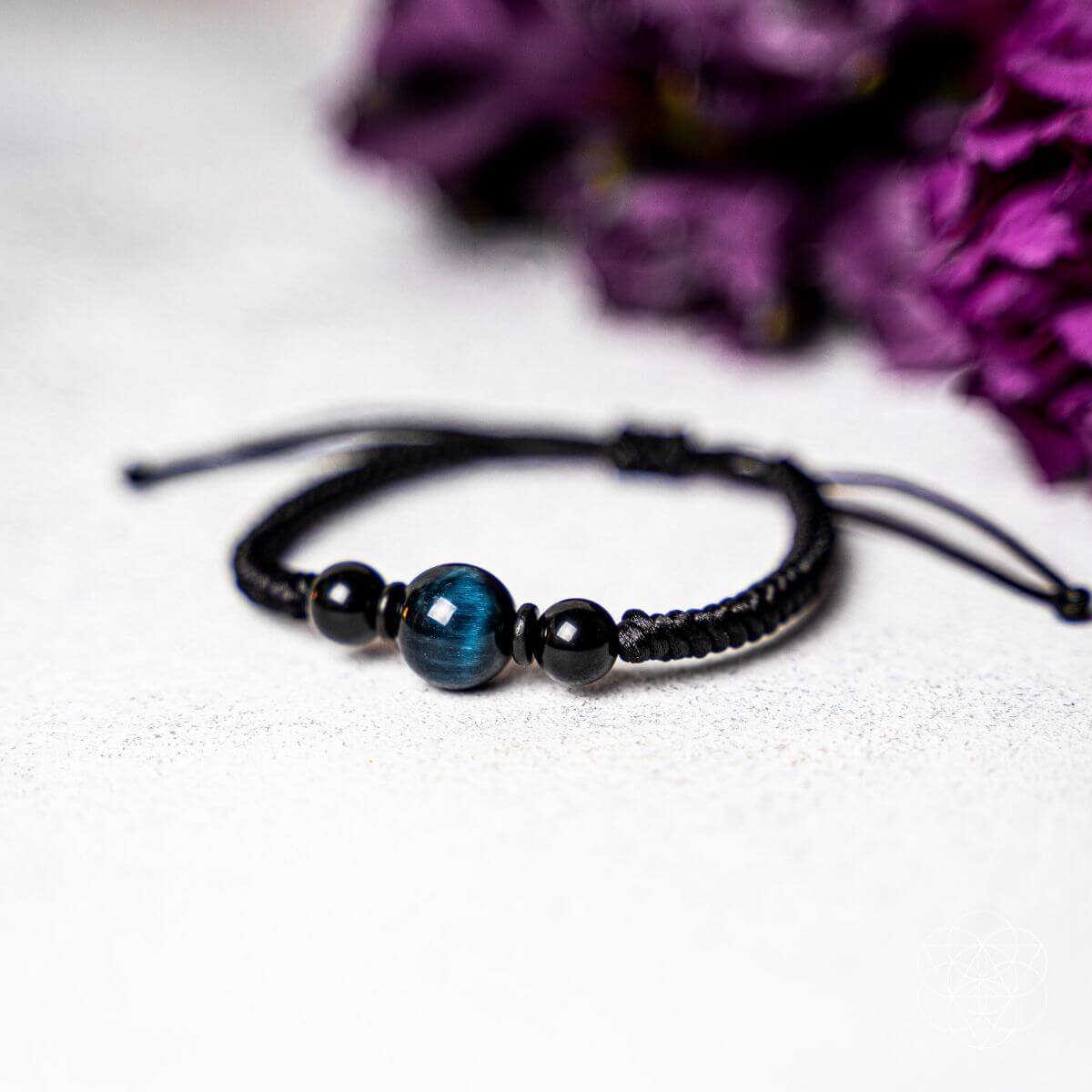 A black beaded bracelet with a central blue-green tiger's eye bead, displayed on a textured light gray surface with blurred purple flowers in the background.