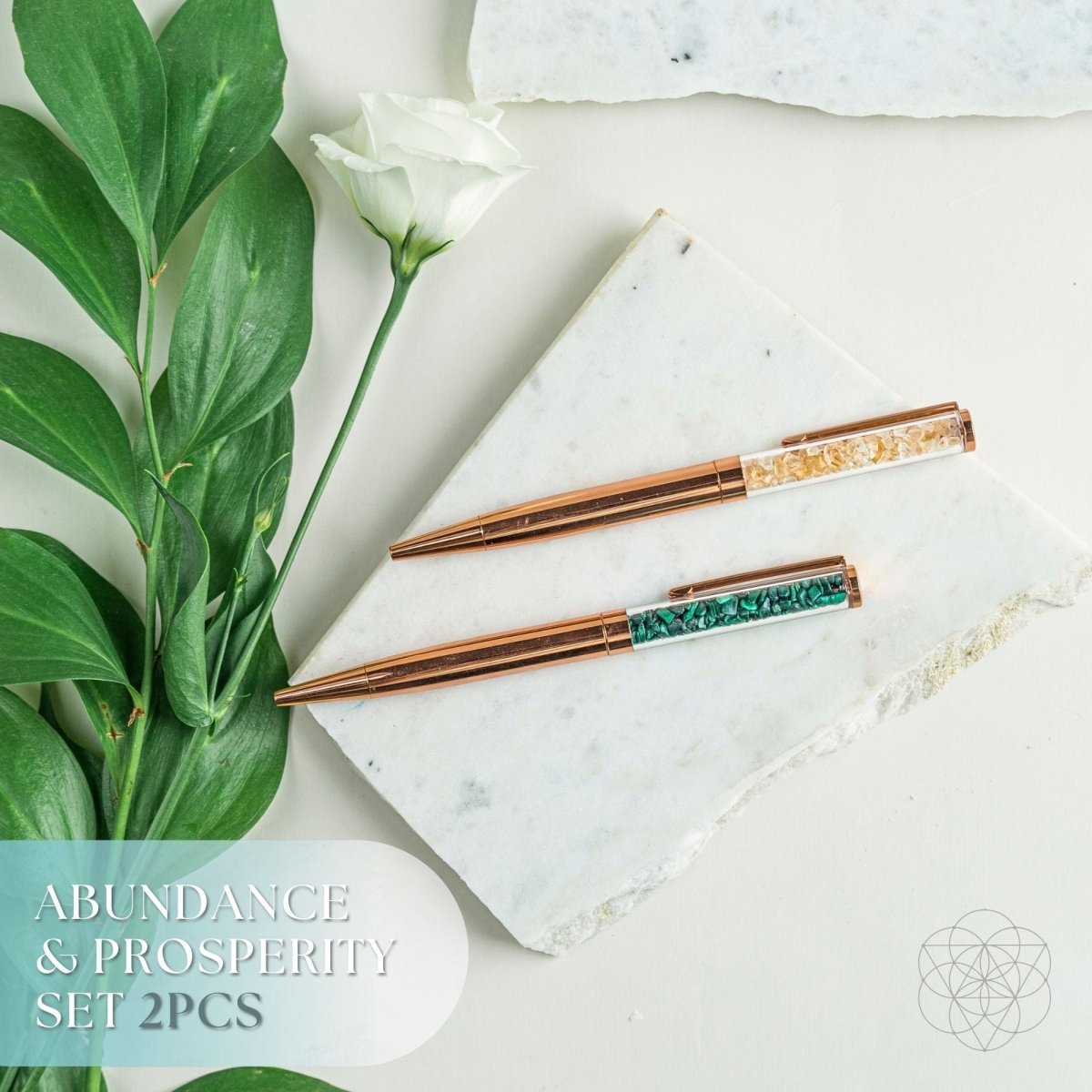 Two rose gold chopsticks with green gemstones on a marble surface with leaves and a flower.