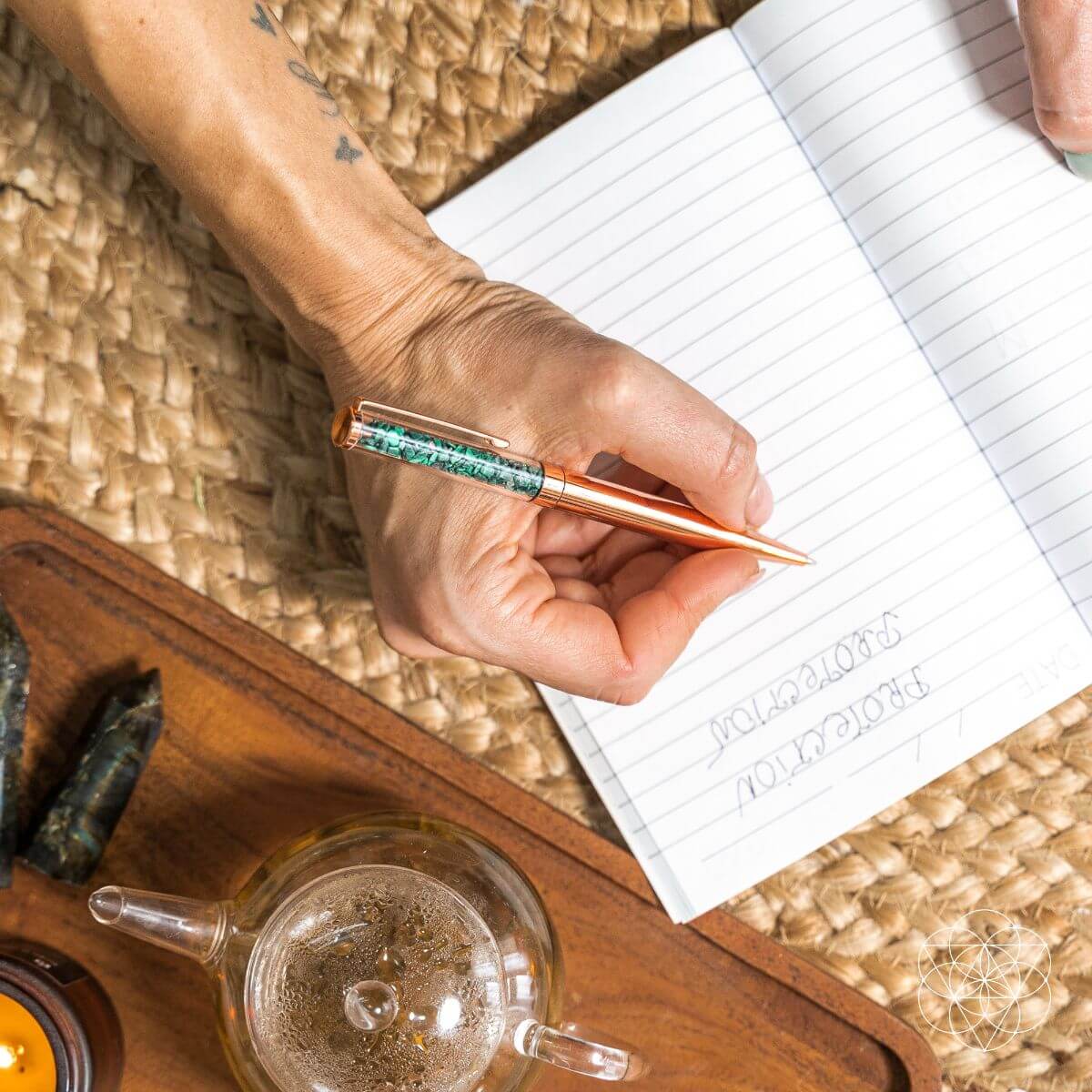 Hand holding a pen over a notebook with a teacup and saucer in the foreground.